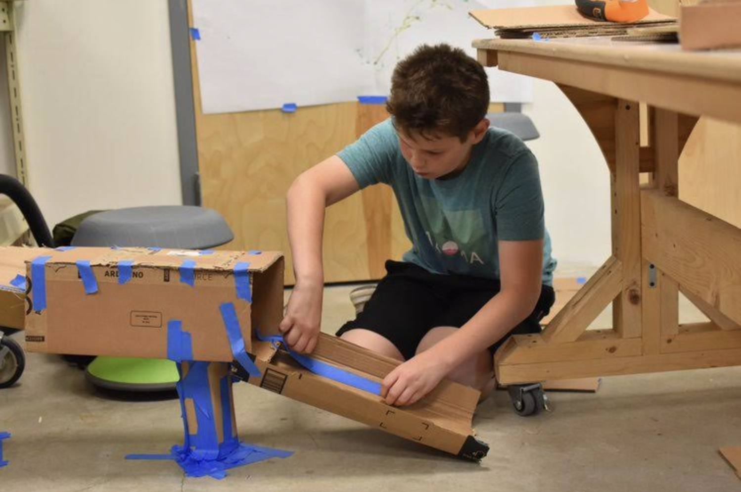 A young person is deeply engaged in building a structure with cardboard and blue tape. They are seated on the floor of a workshop area, surrounded by building materials and tools, demonstrating a hands-on approach to learning and creativity at Court Street Arts.